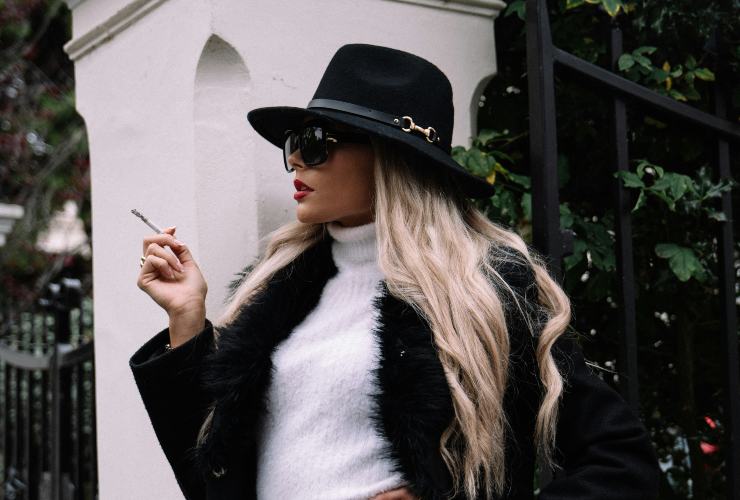 Donna elegante che fuma una sigaretta. Posa con outfit cappotto e cappello nero. Dolcevita bianco
